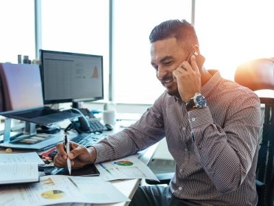 Man on a cell while working at a desk with computers and papers on the desk.