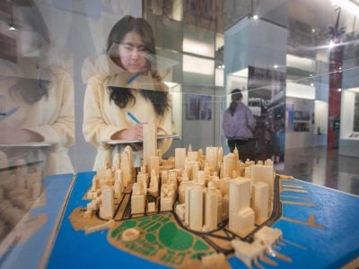 Female student takes notes while observing a wooden architectural model of a city displayed in a glass case. The background features an exhibition space with displays, informational panels.