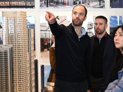 A man gestures toward an architectural display while standing beside a scale model of a high-rise building, as several people observe in a design showroom.