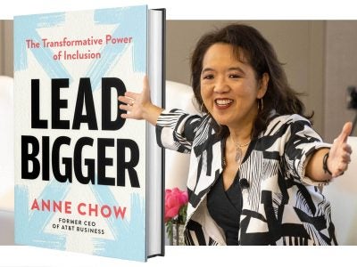 A woman in a black and white blazer with her hands outstretched to the right of an enlarged image of the book she authored.