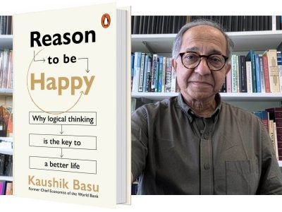 Man stands in front of a book shelf and to his left is an enlarged image of the book he authored.