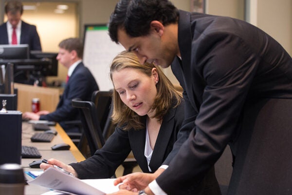 A woman in business formal attire at a desk, holding study materials, a man in business formal attire next to her in conversation