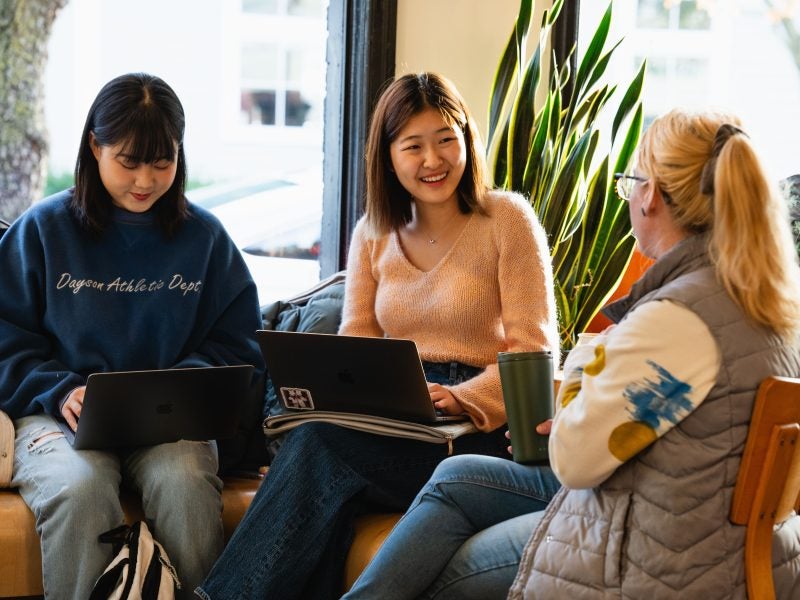 Two students with laptops sit near a window in a coffee shop and interview the manager.