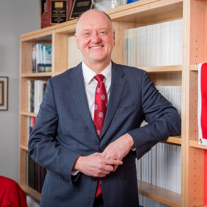 Dean Andrew Karolyi smiling, leaning against bookshelf in his office