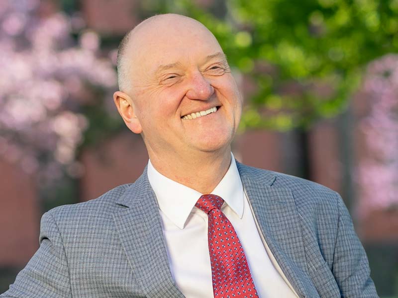 Andrew Karolyi sits outdoors in front of blooming trees.