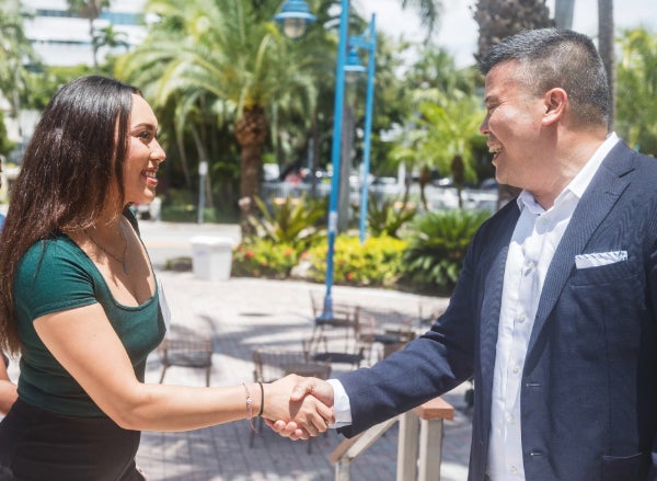 A student shakes the hand of an alum outside with palm trees in background.