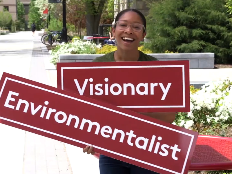 Video thumbnail image of a young woman in front of Warren Hall holding a sign with the word 'Visionary' and a sign with the word 'Environmentalist' while smiling