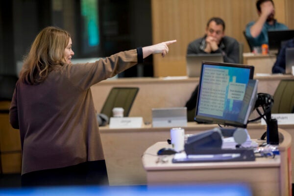 A professor explains a topic at the front of a full lecture hall classroom by pointing her finger and speaking in front of a screen with attentive students looking on