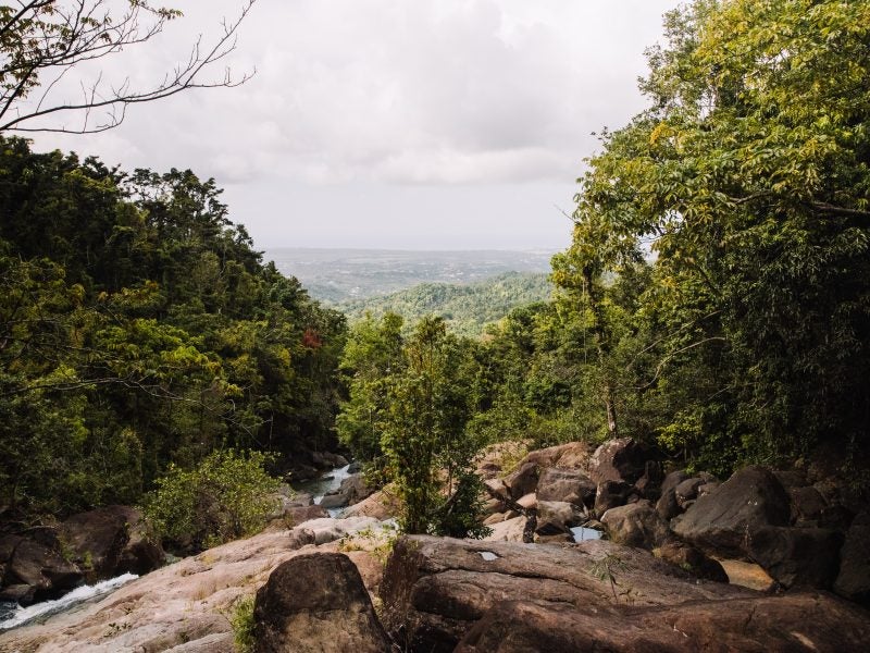 A rocky trail and river with rolling green hills in the background of El Yunque.