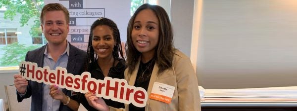 Nolan School undergraduate students smile in business casual attire while holding a cutout sign spelling out the hashtag #HoteliesforHire