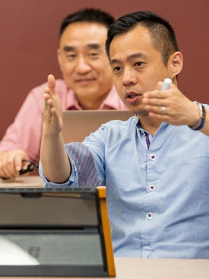 H1-B student gestures while explaining something at a desk in a classroom setting