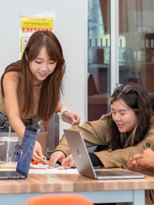 Two Cornell students working together on a laptop at a table, one leaning in to help the other