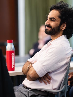 H1-B students sit with his arms crossed, smiling, with a Cornell water bottle on the desk beside him