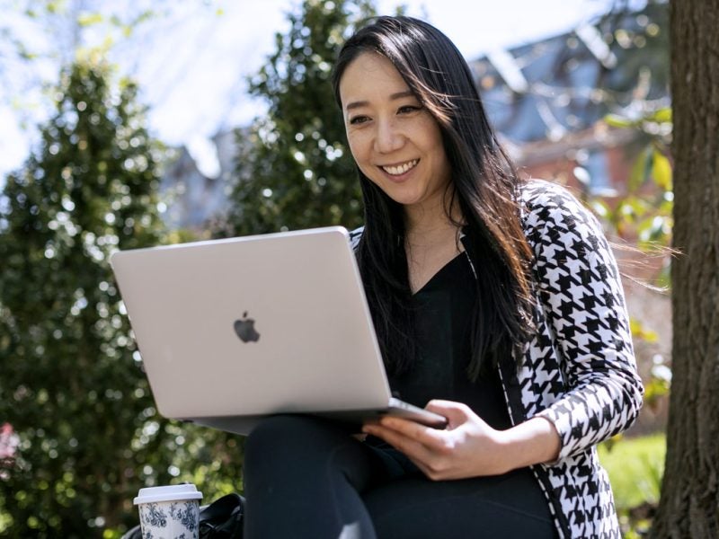 A young woman enrolled in the Cornell MBA program works on her laptop outdoors among grass and trees while smiling