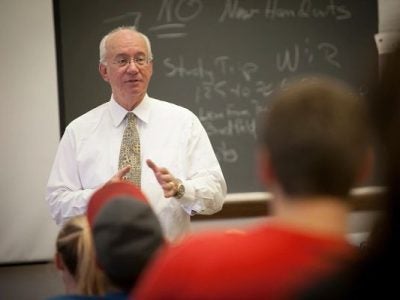 WAyne Knoblauch stands in front of a chalkboard in a white shirt and tie facing students seated in class.