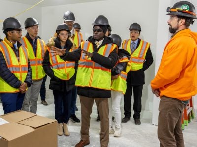 Students, wearing hard hats and construction vests, gather in an unfinished apartment; the student in the center is speaking.
