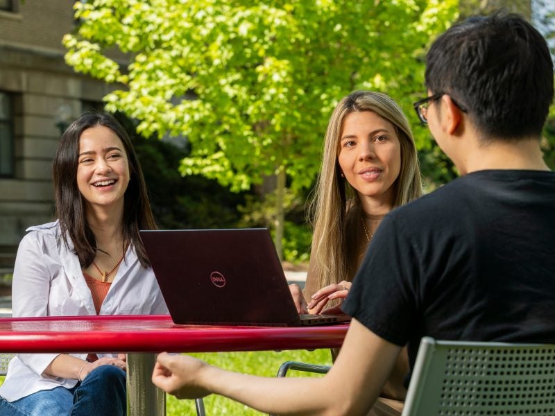 Two young women and one young man, students in Cornell Dyson School's MPS in Applied Economics and Management program, smile and talk at an outdoor table with a green-leaved tree in the background