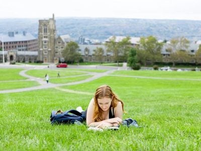 A student lays on her belly reading on the grass, with Cornell's West Campus in the background.