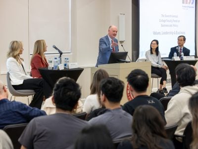 Dean Andrew Karolyi speaks at a podium with arm outstretched, while four panelists and the audience look on.