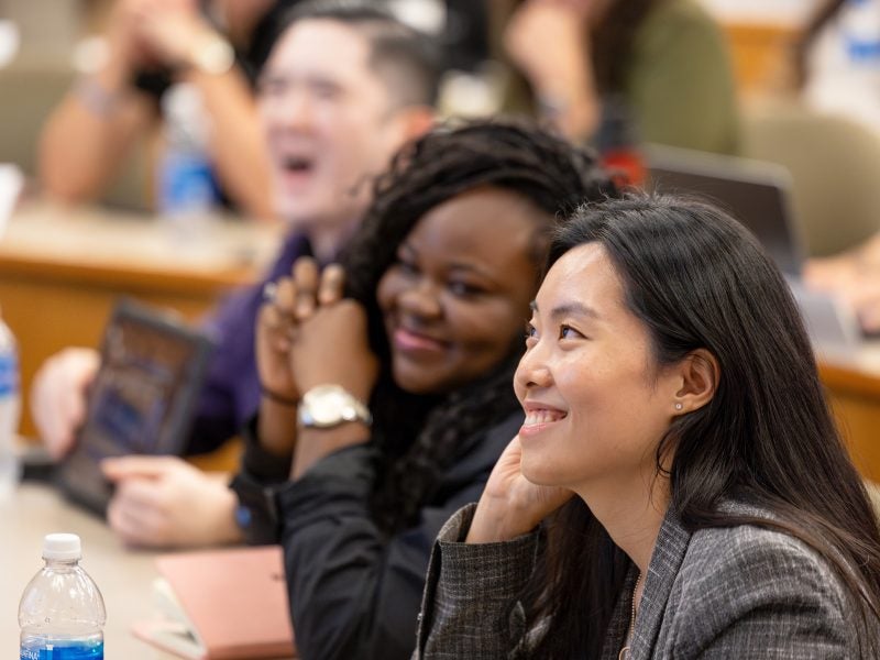 Young professional students from Cornell's M.S. in Business Analytics program, dressed in business casual atire, smile while during class in a lecture hall