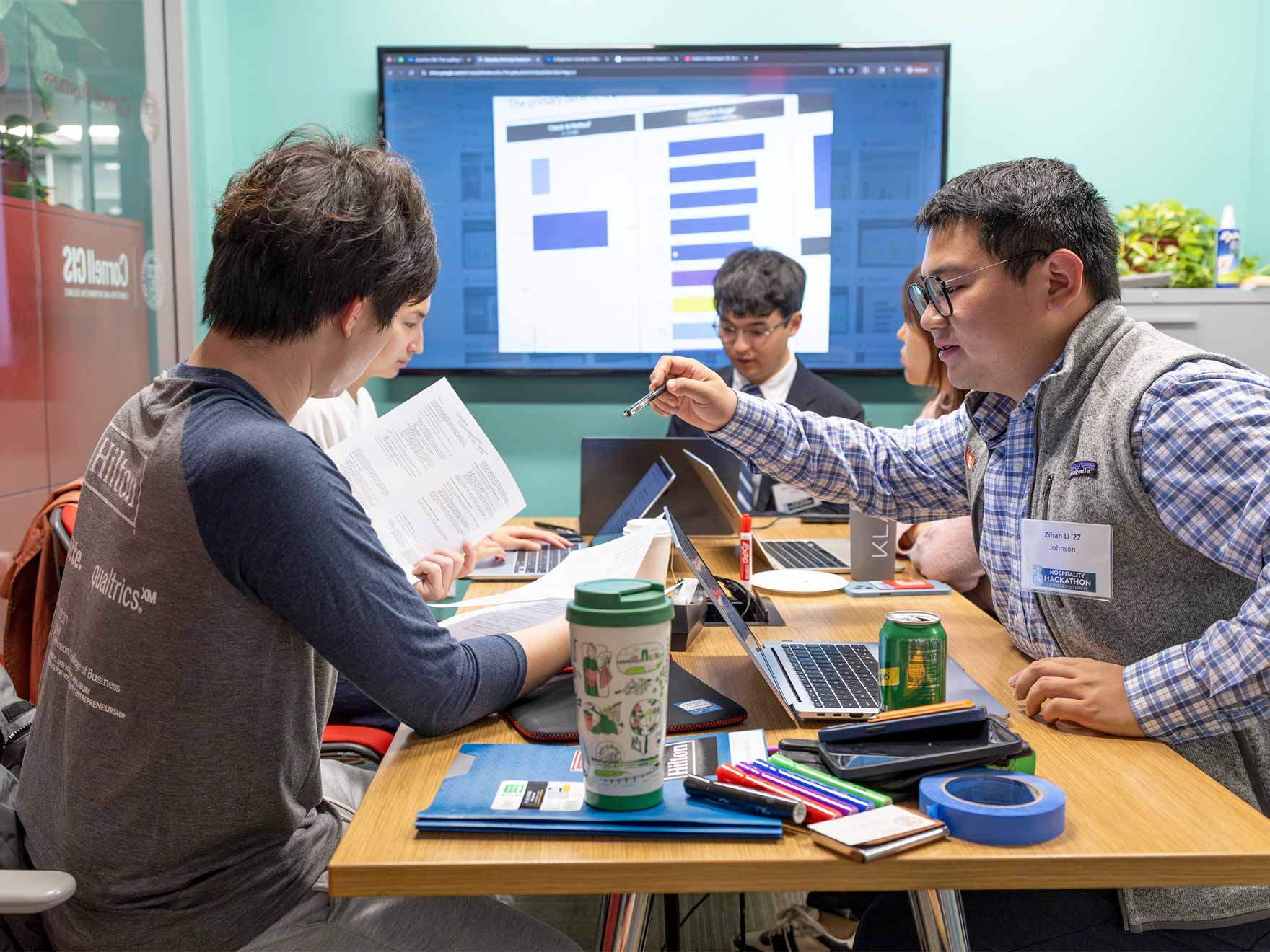 Five students work at a table with a large screen on the back wall of the room.