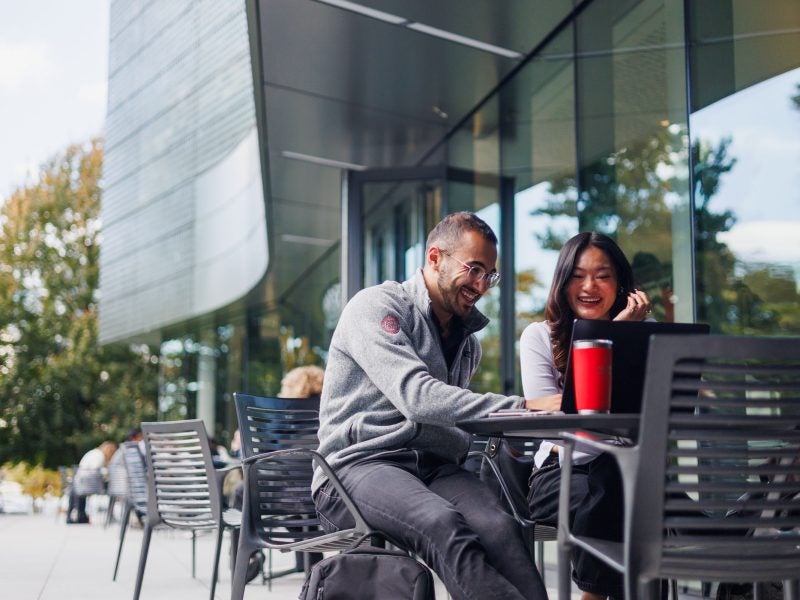 A man and a woman, who are enrolled in the Johnson Cornell Tech MBA program, both smiling, meet at an outdoors cafe outside a contemporary glass building on Roosevelt Island NYC to discuss an assignment