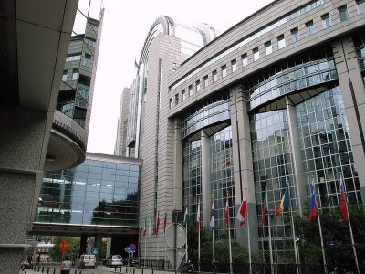 A picture of a white and mirrored building with European Union countries’ flags on its façade, taken from outdoors at the ground level.