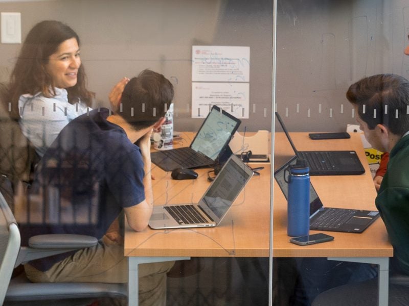 Cornell MBA students sit at a table to collaborate on a project in a glass-walled room, using their laptops