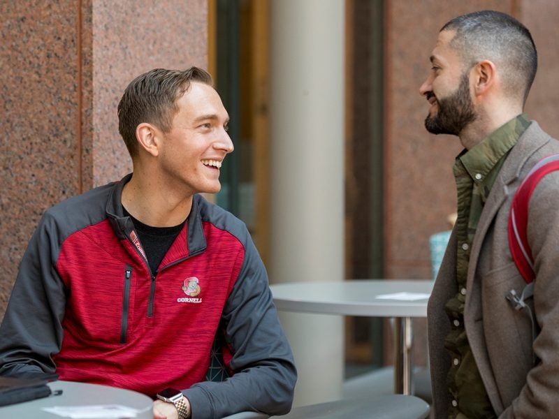 Two men in business casual attire smile while discussing coursework as part of the Cornell MBA's Strategy & Consulting Immersion