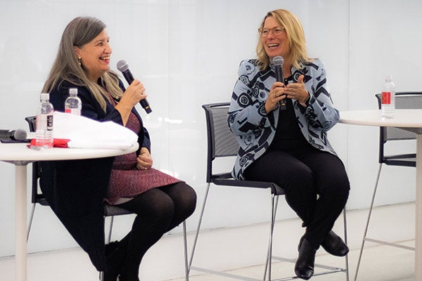 2 women seated on stool-height chairs at the front of a lecture hall, holding microphones, talking, and laughing.