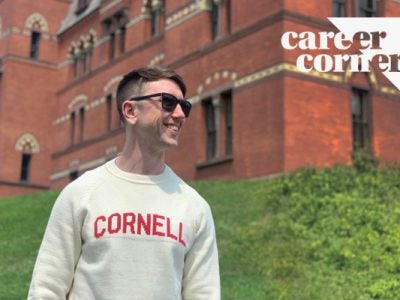 a young man wearing a Cornell sweatshirt and sunglasses standing in front of Sage Hall and smiling.