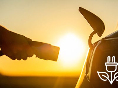 photo of a hand holding an electric charger and about to insert it into the side of a car with a burst of light from the rising sun in the gap between the charger and the car