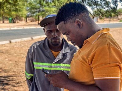 Two men look at a mobile phone near a road in a dry landscape.