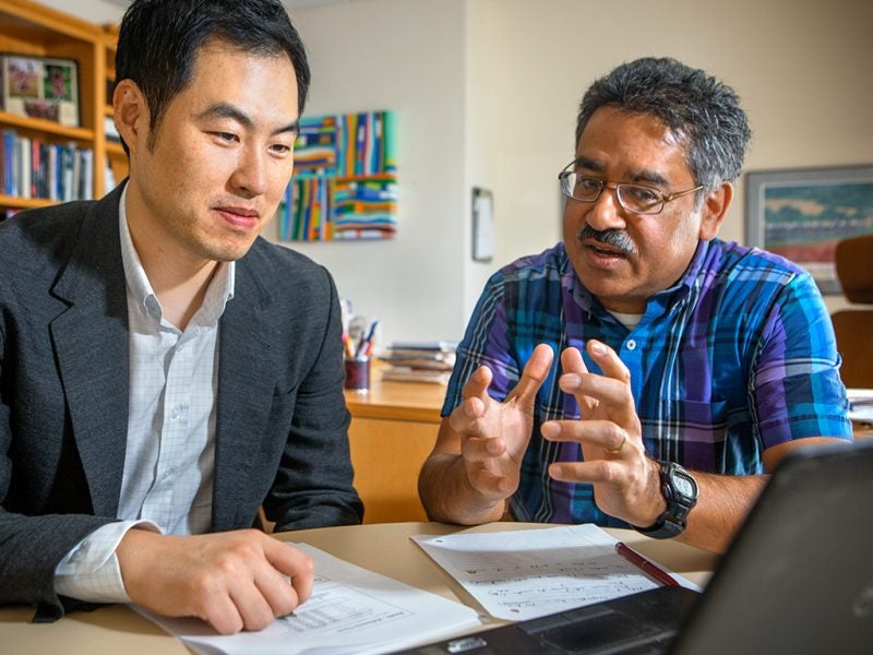 A young man in business casual attire sits at a desk with documents while a professor speaks to him as part of Cornell's Ph.D. in Management.
