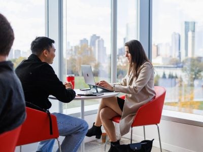 Two Cornell students collaborating at a small table near floor-to-ceiling windows with a city skyline behind them