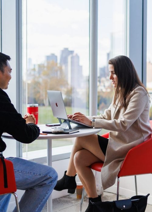 Two Cornell students collaborating at a small table near floor-to-ceiling windows with a city skyline behind them