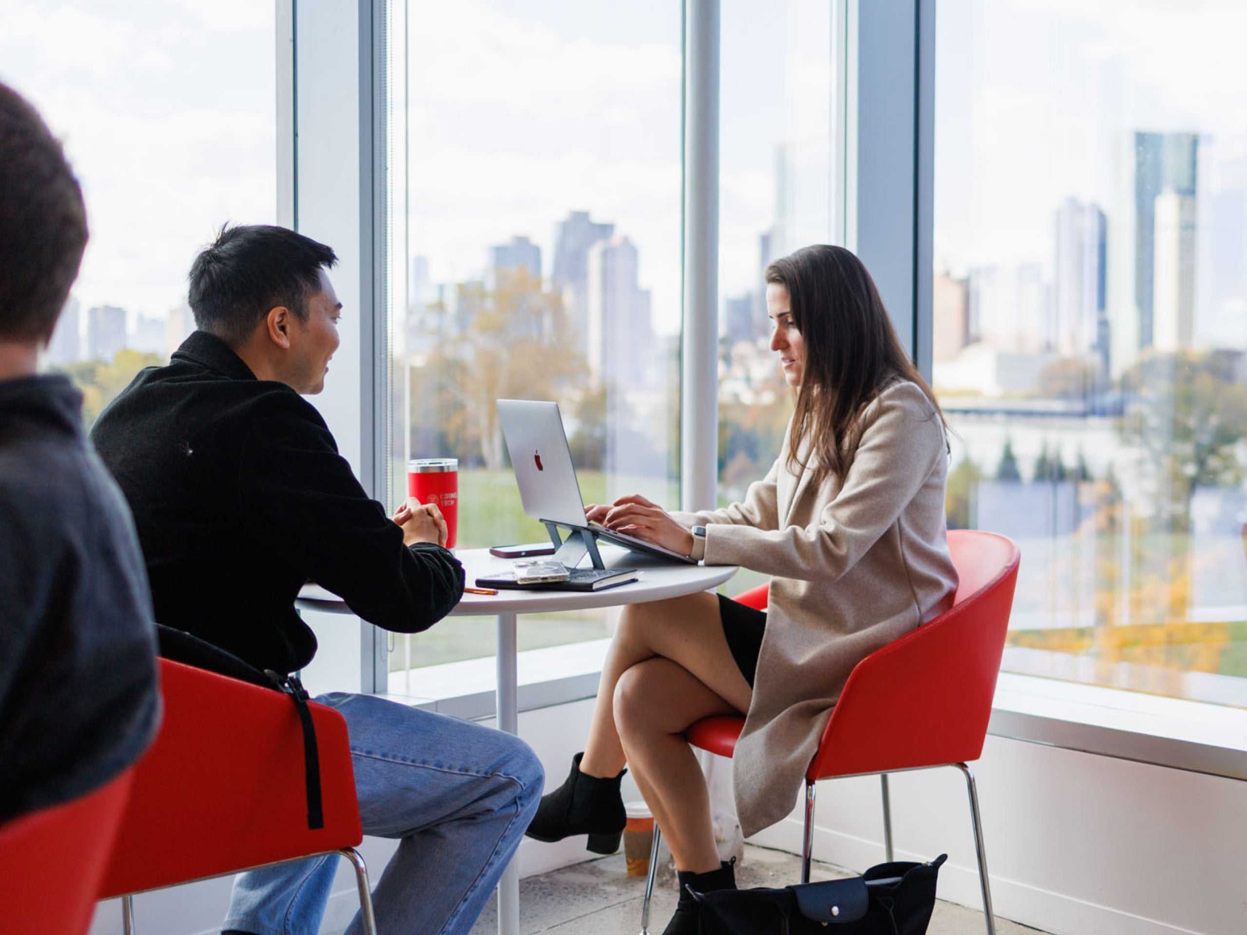 Two Cornell students collaborating at a small table near floor-to-ceiling windows with a city skyline behind them