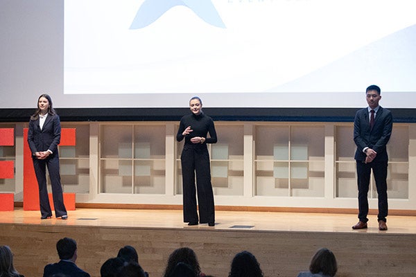 Two women and one man stand on a stage as the woman standing in the middle gestures and addresses the audience.