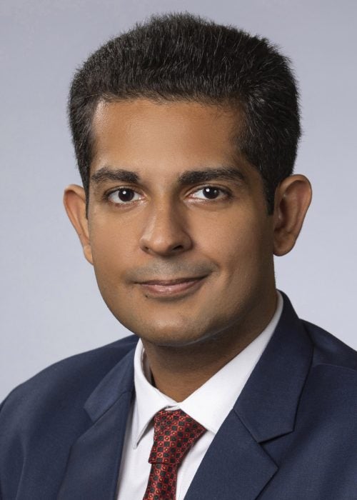 Aditya Sindhu, MBA &rsquo;24, a Cornell MBA student dressed in business formal attire, smiles at the camera in a studio portrait featuring a bluish gray background