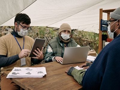 one woman and two men sitting outside at a table conferring and working with a laptop and tablets