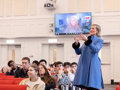 A woman in a lecture hall with students seated looks up at a screen showing another women.