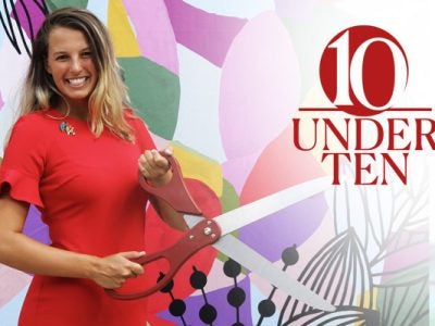a woman standing in front of a brightly colored background holding a huge pair of scissors cutting a ribbon and smiling. the 10 Under 10 graphic identifier is overlaid on the photo.