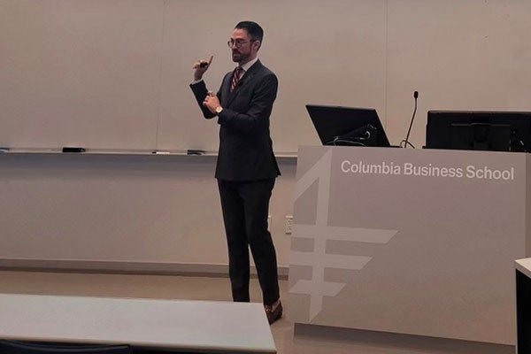 Andrew Foley dressed in a business suit, gesturing and speaking at the front of a lecture hall.