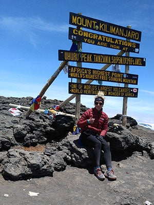 Anissa Buckley sitting in front of a sign that reads: Mount Kilimanjaro, Congratulations you are now at Uhuru Peak, Tanzania, 5895 M / 19341 Ft Africa&rsquo;s highest point, World&rsquo;s highest free-standing mountain