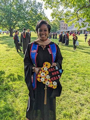 a woman in a graduation gown holding her graduation cap decorated with the words &ldquo;MBA Mom&rdquo; and flowers, standing on the Cornell Arts Quad. 