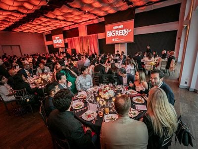 a large banquet hall filled with people seated at round tables and with two large red banners with the text Big Red Bash hanging on either side of the stage.