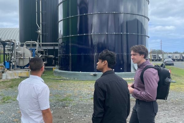 Two team members are talking with an operating engineer wearing a white polo and glasses in front of two large containment vats connected by valve tubes. The engineer is facing the vats and the two team members are facing the engineer.