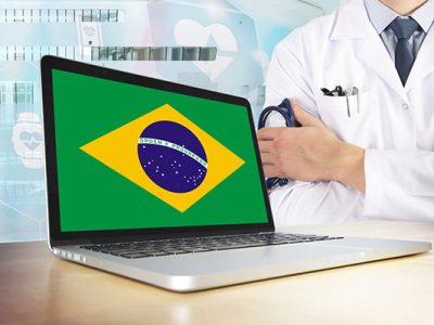 photo of laptop with the Brazilian flag on the screen and in the background, just the right shoulder and left hand of a medical professional in a white lab coat, shirt and tie, holding a stehoscope