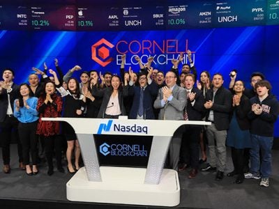 Group photo of Cornell Blockchain members cheering and standing behind the Nasdaq podium at the opening bell, with Cornell Blockchain on the wall behind them and on the podium in front of them