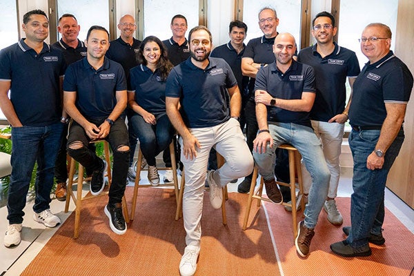 A group of 12 people, standing or seated on stools and smiling, wearing matching navy blue &ldquo;Team COVU&rdquo; polo shirts. 